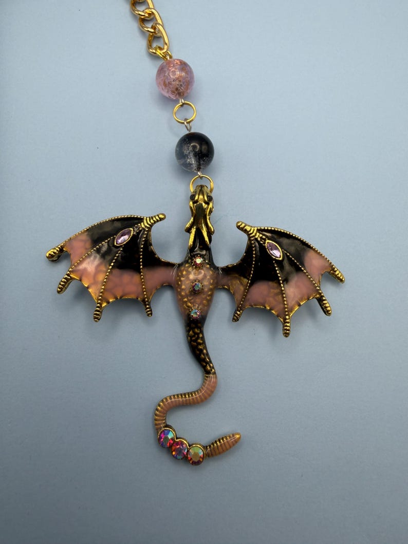 May include: A gold-toned dragon pendant with black and pink wings, adorned with purple and iridescent gemstones. The dragon's body is a mix of pink and brown, with a curled tail. The pendant hangs from a gold chain with pink and black beads.