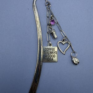 May include: Silver metal bookmark with hanging charms. Charms include a purple gem, a coffee cup, a heart, and a coffee bean. A rectangular charm reads "COFFEE IS ALWAYS A GOOD IDEA". The bookmark has a curved top and a slender design.