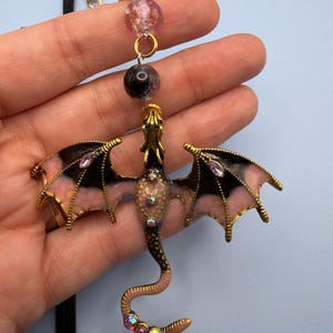 May include: A gold-toned dragon pendant with black and pink wings, adorned with iridescent rhinestones. The dragon's body is a light pink color. The pendant hangs from a gold chain with pink and black beads.