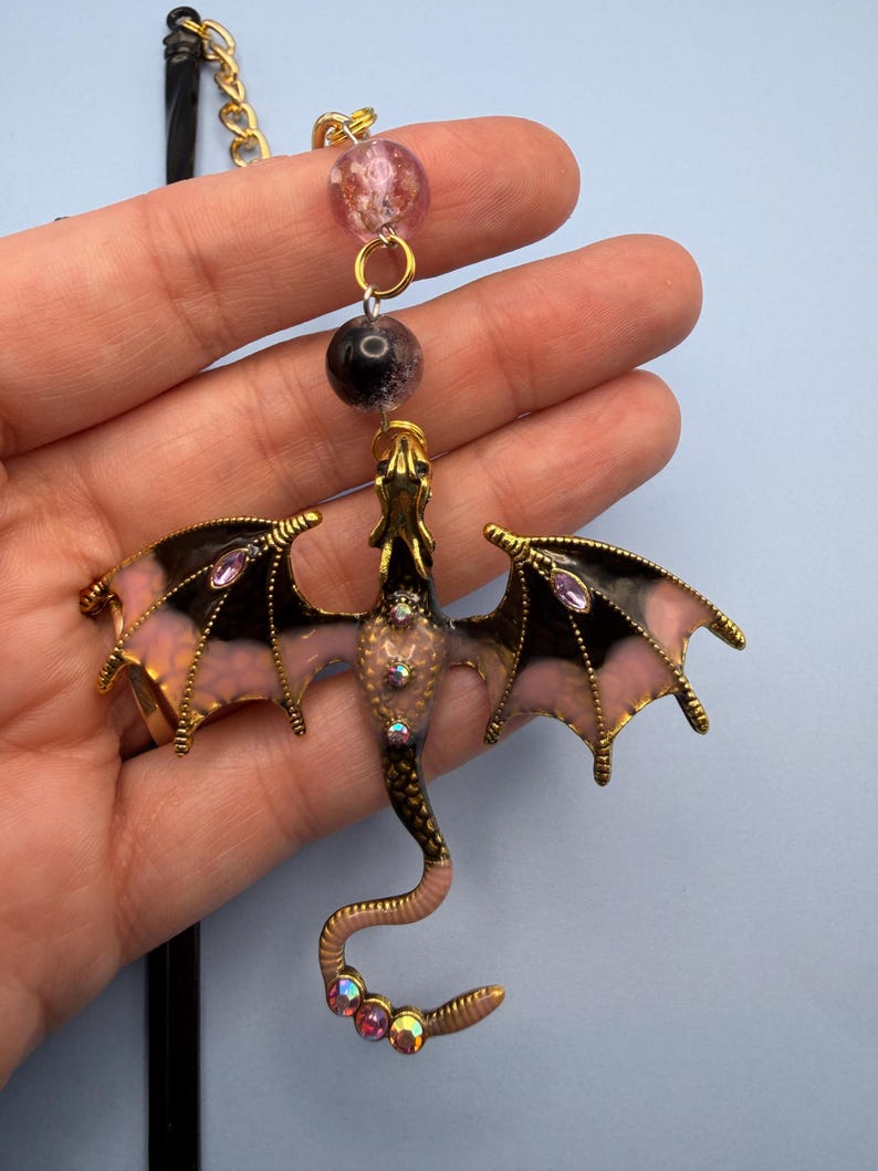 May include: A gold-toned dragon pendant with black and pink wings, adorned with iridescent rhinestones. The dragon's body is a light pink color. The pendant hangs from a gold chain with pink and black beads.
