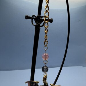May include: A decorative dragon pendant with a gold chain and black sword-shaped stand. The dragon has outstretched wings and is brown with red accents. Two beads are suspended between the chain and the dragon.