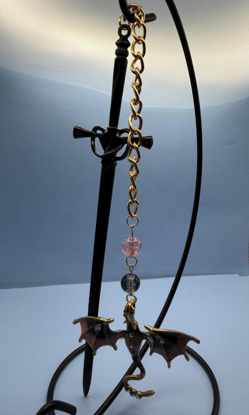 May include: A decorative dragon pendant with a gold chain and black sword-shaped stand. The dragon has outstretched wings and is brown with red accents. Two beads are suspended between the chain and the dragon.
