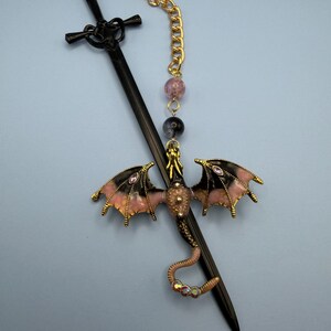 May include: A black sword-shaped hair stick with a decorative dragon charm. The dragon has pink and black wings, gold accents, and purple gems. A gold chain and beaded accents complete the design.