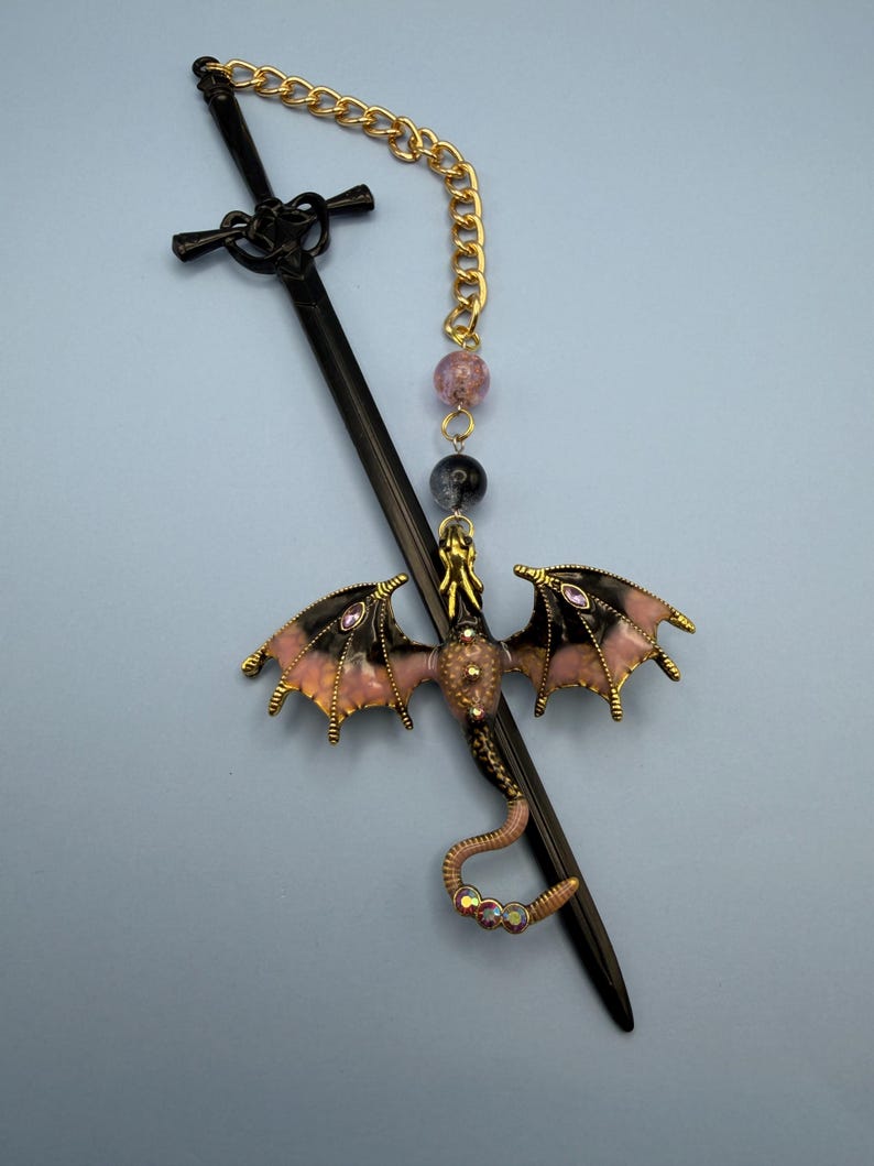 May include: A black sword-shaped hair stick with a decorative dragon charm. The dragon has pink and black wings, gold accents, and purple gems. A gold chain and beaded accents complete the design.