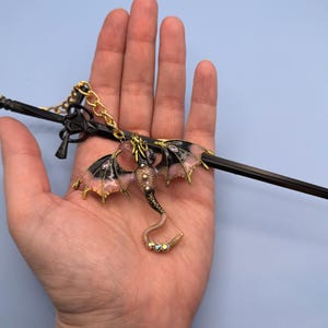 May include: A black metal sword-shaped hair accessory with a dragon charm. The dragon has pink and black wings with gold accents, a gold chain, and a beaded tail. The accessory is held in a hand against a light blue background.