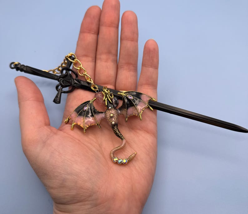 May include: A black metal sword-shaped hair accessory with a dragon charm. The dragon has pink and black wings with gold accents, a gold chain, and a beaded tail. The accessory is held in a hand against a light blue background.