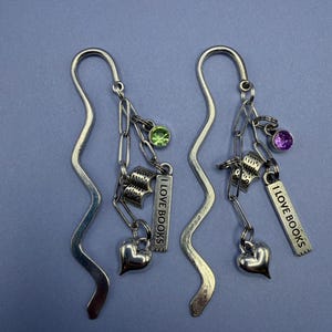 Puede incluir: Dos marcadores de metal plateado con un diseño ondulado. Cada marcador tiene una cadena con un pequeño dije de libro abierto, un dije de corazón, una gema verde o morada y una etiqueta rectangular que dice "I LOVE BOOKS."