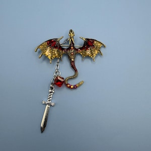 Puede incluir: Broche con forma de dragón con alas extendidas, con detalles rojos y dorados. Una pequeña espada y una gema roja cuelgan del dragón. El cuerpo del dragón es rojo y dorado, con un diseño detallado. El fondo es azul claro.