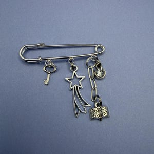 May include: A silver-toned safety pin with dangling charms. Charms include a key, a shooting star, a book, and a clear crystal. The pin is set against a solid blue background.