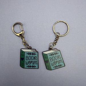 Puede incluir: Dos llaveros con herrajes dorados. Cada llavero presenta un dije de libro de color verde azulado con las palabras "BOOK WORM" y un corazón. Los dijes tienen un diseño de hojas decorativas. El fondo es de color gris sólido.
