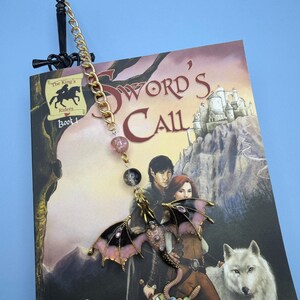 May include: A fantasy-themed bookmark featuring a black sword design at the top, a gold chain, and a dragon charm. The dragon has pink and black wings and is adorned with rhinestones. The bookmark is placed in a book titled "Sword's Call".