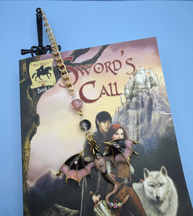 May include: A fantasy-themed bookmark featuring a black sword design at the top, a gold chain, and a dragon charm. The dragon has pink and black wings and is adorned with rhinestones. The bookmark is placed in a book titled "Sword's Call".