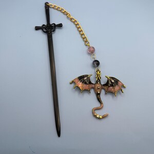 May include: A black sword-shaped bookmark with a decorative handle is next to a dragon-shaped charm. The dragon charm has pink and black wings, a gold body, and is connected to a gold chain with beads.