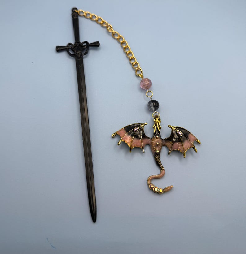May include: A black sword-shaped bookmark with a decorative handle is next to a dragon-shaped charm. The dragon charm has pink and black wings, a gold body, and is connected to a gold chain with beads.