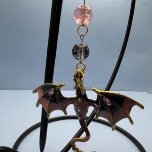 May include: A decorative dragon ornament with outstretched wings, featuring a gold-colored head and body, and black and pink wings. The dragon is adorned with iridescent gems and hangs from a chain with pink and clear beads.