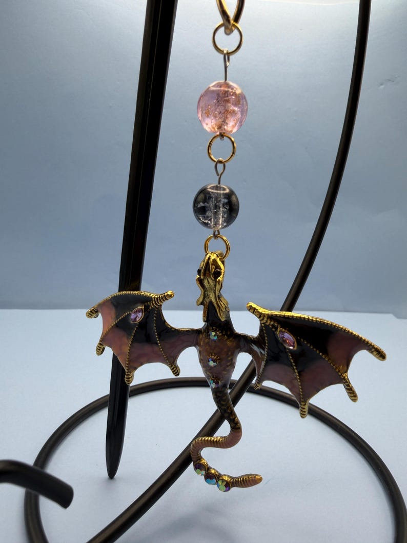 May include: A decorative dragon ornament with outstretched wings, featuring a gold-colored head and body, and black and pink wings. The dragon is adorned with iridescent gems and hangs from a chain with pink and clear beads.