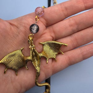 May include: A gold-colored dragon pendant with detailed wings and body, suspended from a black cord and gold chain. The pendant is connected to two decorative beads, one pink and one black. The dragon is held in a person's hand against a light blue background.