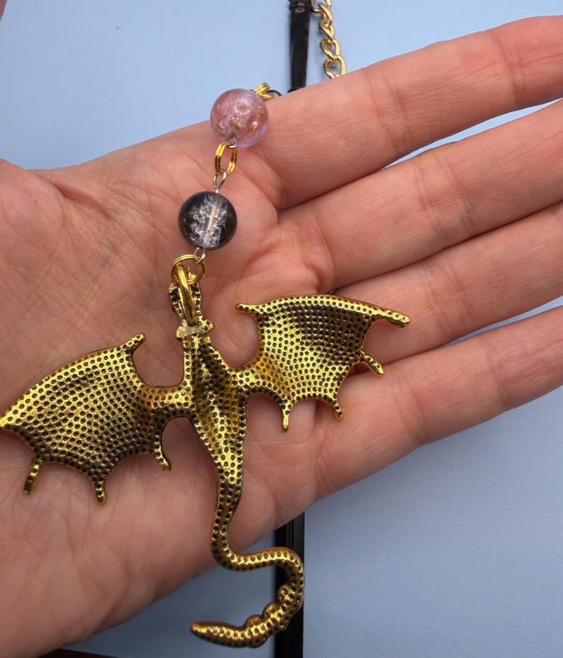 May include: A gold-colored dragon pendant with detailed wings and body, suspended from a black cord and gold chain. The pendant is connected to two decorative beads, one pink and one black. The dragon is held in a person's hand against a light blue background.
