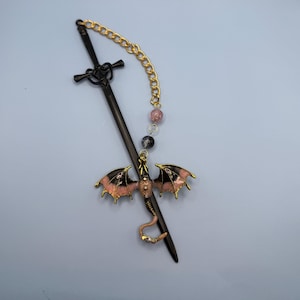 May include: A black sword-shaped hair stick with a gold chain and pink and black dragon charm. The dragon has pink and black wings with gold trim. The chain has pink and black beads.