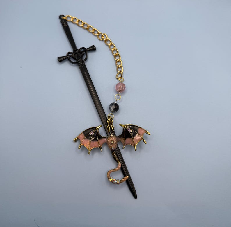 May include: A black sword-shaped hair stick with a gold chain and pink and black dragon charm. The dragon has pink and black wings with gold trim. The chain has pink and black beads.