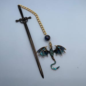May include: A dark metal hair stick shaped like a sword, connected by a gold-toned chain to a dark blue and bronze dragon charm. The dragon features iridescent blue wing accents and small blue crystals along its tail, with a dark blue bead.