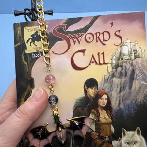 May include: A fantasy novel titled "Sword's Call" with a decorative bookmark. The bookmark features a black sword design with a gold chain, pink and black beads, and a dragon charm with pink and purple accents.