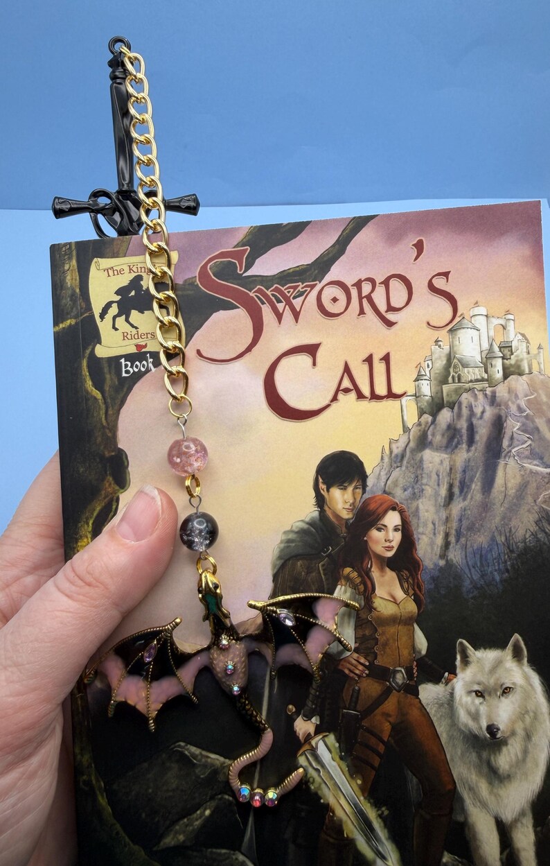 May include: A fantasy novel titled "Sword's Call" with a decorative bookmark. The bookmark features a black sword design with a gold chain, pink and black beads, and a dragon charm with pink and purple accents.
