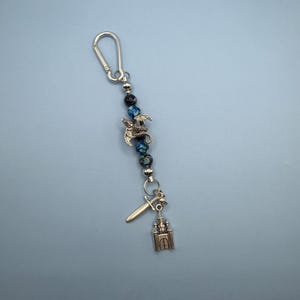 May include: A keychain featuring a silver carabiner clip, blue and black speckled beads, a silver dragon charm, a sword charm, and a castle charm. The keychain is designed with fantasy and medieval themes.