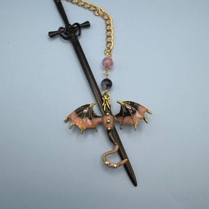 May include: A black sword-shaped hair stick with a gold chain and a dragon charm. The dragon has pink and black wings, and a gold body. The chain has pink and black beads.
