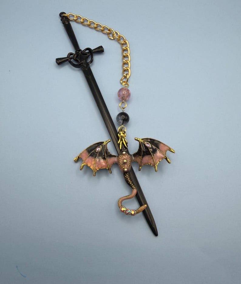 May include: A black sword-shaped hair stick with a gold chain and a dragon charm. The dragon has pink and black wings, and a gold body. The chain has pink and black beads.
