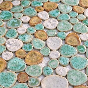 Puede incluir: Una hoja de azulejos de mosaico con piezas de cerámica lisas, redondeadas y con forma de guijarro en tonos verde espuma de mar, blanco y ámbar. Cada azulejo muestra un patrón de esmalte cristalino único, creando una superficie texturizada brillante.