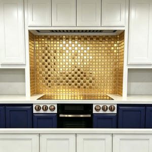 May include: A kitchen stove area featuring a metallic gold square tile backsplash, a black glass cooktop, and a black oven. The unit is flanked by dark blue lower cabinets and white upper cabinets with matching gold-toned control knobs.