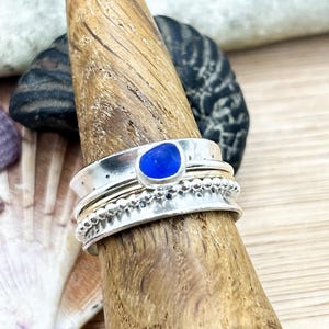 Pode incluir: Um anel de prata com uma pedra azul é exibido em um suporte de anel de madeira. O anel tem uma banda texturizada e uma banda menor com contas. O fundo inclui conchas e pedras.