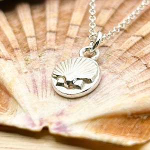 May include: A silver necklace featuring a small, round pendant with a raised seashell design. The pendant hangs from a delicate silver chain, showcasing a polished metallic finish that reflects light, creating a bright and clear appearance.
