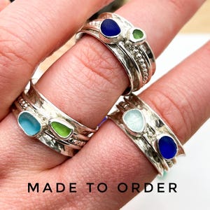Puede incluir: Primer plano de varios anillos de plata, cada uno con una piedra de vidrio marino única en tonos de azul, verde y aguamarina. Los anillos tienen una banda texturizada y se muestran en los dedos. El texto "MADE TO ORDER" está en la parte inferior.