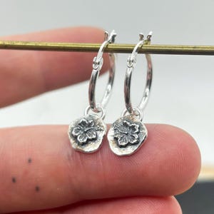 May include: Silver hoop earrings with a floral charm. The charms feature a flower design. The earrings are shown against a plain background.