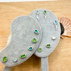 Può includere: Una collezione di orecchini a lobo con piccoli pezzi lisci di vetro marino nelle tonalità del verde, verde acqua e bianco, montati su argento. Gli orecchini sono esposti su due supporti in velluto grigio a forma di rene con perni.