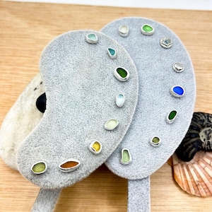 Può includere: Una collezione di orecchini a bottone in argento sterling con vetro marino colorato di varie forme e dimensioni. Gli orecchini sono esposti su espositori per orecchini in velluto grigio. I colori del vetro marino includono verde, blu, giallo e marrone.