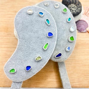Può includere: Due espositori per orecchini in velluto grigio a forma di fagiolo, ciascuno con diverse paia di orecchini a lobo. Gli orecchini sono composti da piccoli pezzi di vetro marino levigato e liscio nei toni del blu, del verde e del bianco.