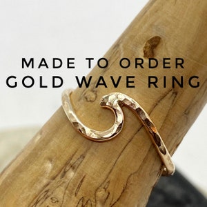 May include: A gold wave ring with a hammered texture is displayed on a wooden dowel. The ring's design mimics a curling wave. The text "MADE TO ORDER GOLD WAVE RING" is above the ring.