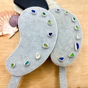 May include: Two gray velvet earring display cards shaped like kidney beans, each featuring multiple small, colorful sea glass studs in shades of blue, green, and white, set in silver-toned metal bezels arranged along the curved edges.