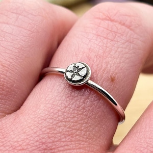 Könnte beinhalten: Ein silberner Ring mit einem kleinen, runden Sanddollar-Anhänger. Der Ring wird an einem Finger getragen.
