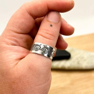 May include: A wide silver band ring featuring a continuous engraved pattern of dark, swirling scrollwork. The metal has a polished finish with recessed black details that highlight the intricate, repeating floral-inspired design elements.