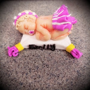 Pode incluir: Uma figurinha de bebê dormindo feita à mão com uma saia listrada rosa e branca, uma faixa de cabeça branca com bolinhas brancas e uma chupeta rosa. O bebê está deitado em um travesseiro branco com o nome "Emily" escrito em preto. O travesseiro tem borlas rosa e amarelas.