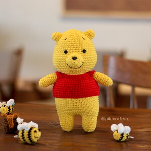 May include: A crocheted Winnie the Pooh plush toy wearing a red shirt, standing on a wooden table with three crocheted bees. The bees are yellow and black with white wings. The caption on the table reads "@pikicraftvn".
