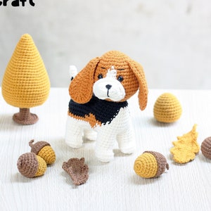 Puede incluir: Un juguete de cachorro beagle de crochet con pelaje marrón, negro y blanco. El juguete está rodeado de bellotas y hojas de crochet en tonos marrones y amarillos. El juguete está sentado sobre una superficie blanca.