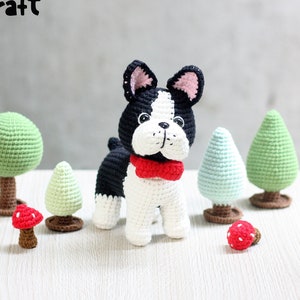 Puede incluir: Un bulldog francés de crochet negro y blanco con una pajarita roja, rodeado de árboles y setas en miniatura de crochet.