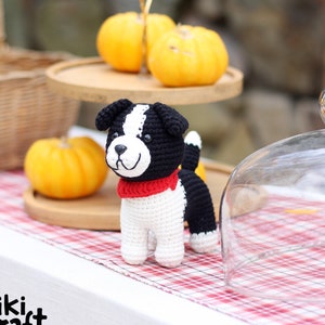 Puede incluir: Un perro de crochet negro y blanco con una bufanda roja. El perro está de pie sobre un mantel a cuadros rojo y blanco. Hay calabazas en el fondo.