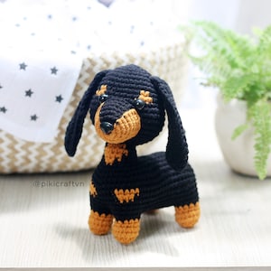 Puede incluir: Un juguete de perro salchicha de crochet negro y marrón con ojos negros y nariz marrón.