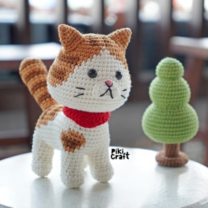May include: A crocheted cat toy with white, brown and orange fur. The cat is wearing a red scarf and has black eyes. The cat is standing on a white surface next to a green crocheted tree. The text "Piki Craft" is visible on the surface.